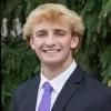 Smiling young man with blond hair wearing a dark suit, white shirt, and purple tie standing outdoors with green foliage background.