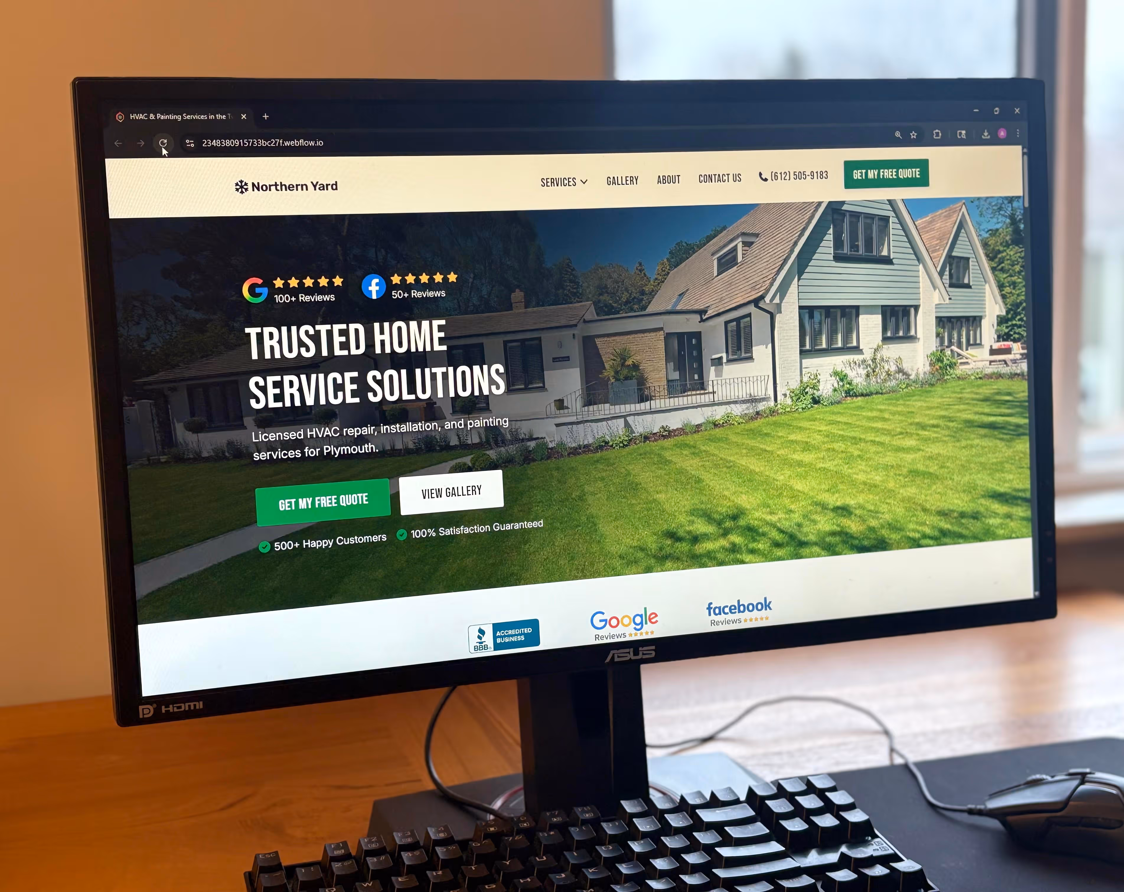 Construction site with workers assembling a gray concrete block retaining wall beside a house, surrounded by trees and gravel.