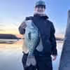 Person wearing dark clothing and a hat holding a large fish near a body of water at sunset.