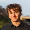 Young man with curly brown hair smiling outdoors with greenery and a clear sky in the background.